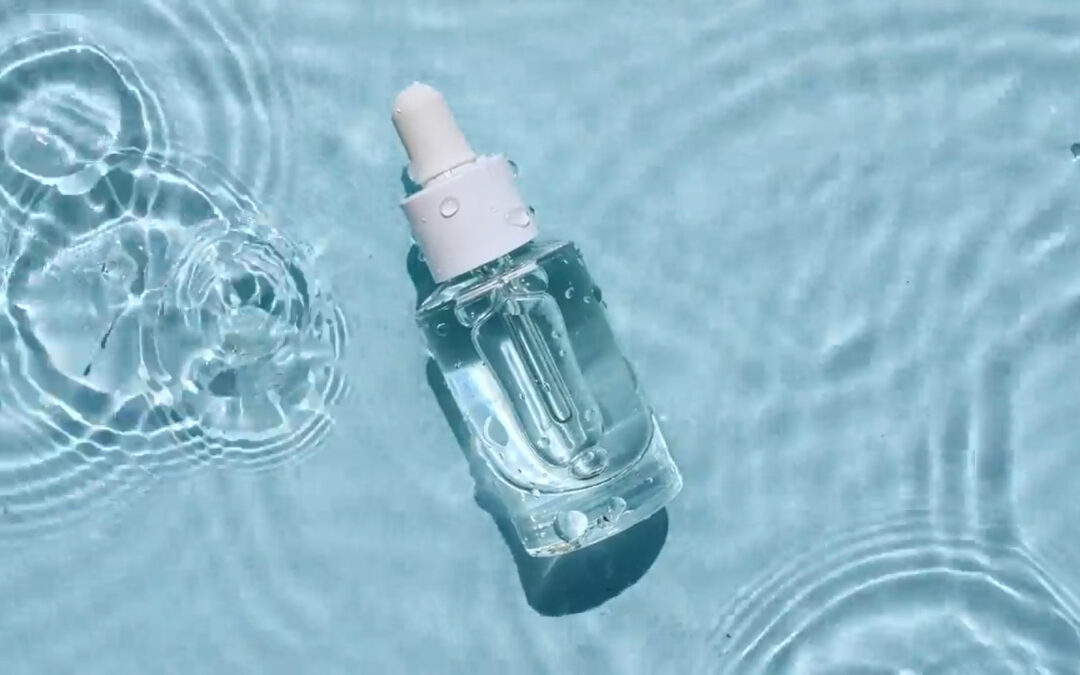 Eine gläserne Kosmetikflasche mit Pipette liegt auf der Wasseroberfläche. Aus der Vogelperspektive ist zu sehen, wie ein Tropfen ins Wasser fällt.